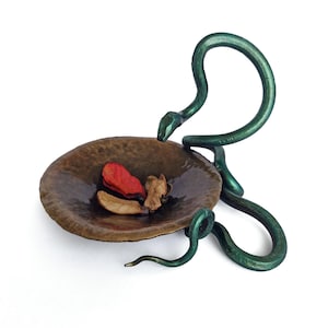 Puede incluir: Una escultura de serpiente de metal verde con un plato redondo de cobre martillado. La serpiente está enroscada alrededor del plato, con la cabeza levantada y la cola enroscada debajo. El plato contiene flores secas y bayas rojas.