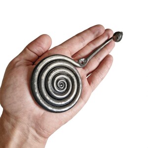 May include: A dark gray metal wall hook featuring a prominent circular spiral design. The hook has a long, slender arm extending from the spiral, ending in a small, rounded knob. A single screw hole is visible near the base of the arm.