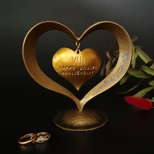 Puede incluir: Una escultura dorada en forma de coraz&oacute;n con un colgante de coraz&oacute;n m&aacute;s peque&ntilde;o con la inscripci&oacute;n "VIII HAPPY BRONZE ANNIVERSARY!" y dos anillos de oro. La escultura tiene un acabado texturizado y est&aacute; sobre una superficie oscura.