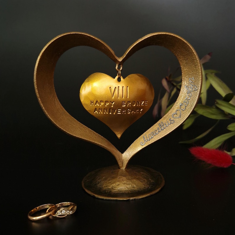 Puede incluir: Una escultura dorada en forma de coraz&oacute;n con un colgante de coraz&oacute;n m&aacute;s peque&ntilde;o con la inscripci&oacute;n "VIII HAPPY BRONZE ANNIVERSARY!" y dos anillos de oro. La escultura tiene un acabado texturizado y est&aacute; sobre una superficie oscura.