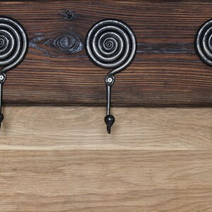 May include: A wall-mounted coat rack featuring three dark gray metal hooks with a prominent spiral design at the top. The hooks are securely attached to a horizontal plank of dark stained wood, which is positioned above a lighter natural wood surface.