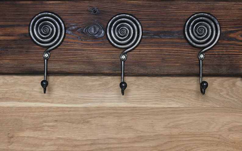 May include: A wall-mounted coat rack featuring three dark gray metal hooks with a prominent spiral design at the top. The hooks are securely attached to a horizontal plank of dark stained wood, which is positioned above a lighter natural wood surface.