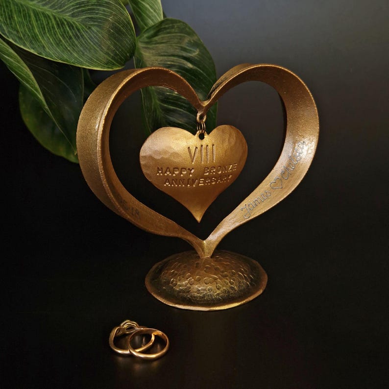 Puede incluir: Una escultura en forma de coraz&oacute;n de color bronce con un colgante de coraz&oacute;n m&aacute;s peque&ntilde;o con la inscripci&oacute;n "VIII HAPPY BRONZE ANNIVERSARY". Dos anillos de oro se encuentran frente a la escultura. La escultura tiene una base texturizada.