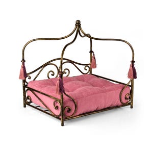 Könnte beinhalten: Dekoratives Haustierbett mit einem rosa Kissen und einem verzierten, antikgoldfarbenen Metallrahmen. Das Bett hat ein Baldachin-Design mit dekorativen Quasten in Rosa- und Lilatönen. Das Bett ist quadratisch.