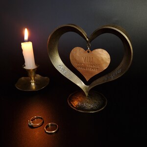 Puede incluir: Una escultura en forma de coraz&oacute;n de bronce con un colgante de coraz&oacute;n m&aacute;s peque&ntilde;o en el interior, con la inscripci&oacute;n "Happy Bronze Anniversary" y n&uacute;meros romanos. La escultura est&aacute; sobre una superficie oscura, con dos anillos y una vela encendida en un portavelas de lat&oacute;n cerca.