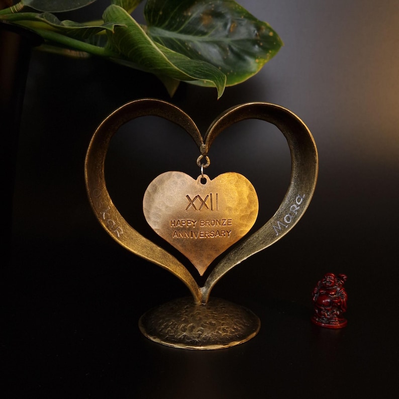 Puede incluir: Un adorno de aniversario en forma de coraz&oacute;n de color bronce. El adorno presenta un marco en forma de coraz&oacute;n m&aacute;s grande con un colgante de coraz&oacute;n m&aacute;s peque&ntilde;o en el centro. El colgante dice "XXII HAPPY BRONZE ANNIVERSARY".