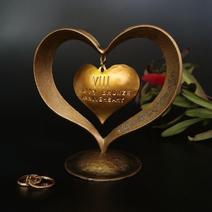 Può includere: Una scultura dorata a forma di cuore per l'anniversario, con un ciondolo a forma di cuore più piccolo con la scritta "VIII Happy Bronze Anniversary". Due fedi nuziali d'oro in primo piano. La scultura è su una superficie scura.