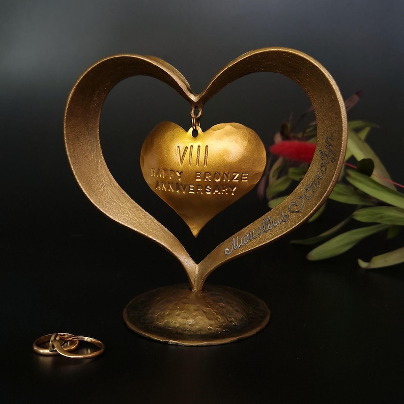 Puede incluir: Una escultura dorada en forma de coraz&oacute;n para aniversario, con un colgante de coraz&oacute;n m&aacute;s peque&ntilde;o que dice "VIII Happy Bronze Anniversary". Dos anillos de boda dorados en primer plano. La escultura est&aacute; sobre una superficie oscura.