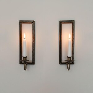 Puede incluir: Dos candelabros de pared con marcos rectangulares y un acabado de bronce envejecido. Cada uno sostiene una vela blanca con una llama encendida. Los candelabros tienen un gancho decorativo debajo del portavelas.