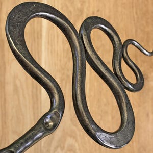 Pu&ograve; includere: Una scultura in metallo forgiato a mano a forma di serpente con una finitura scura e brunita. Il serpente &egrave; raffigurato in una posa sinuosa a forma di S con un corpo liscio e curvo e una testa distinta, dal carattere industriale.