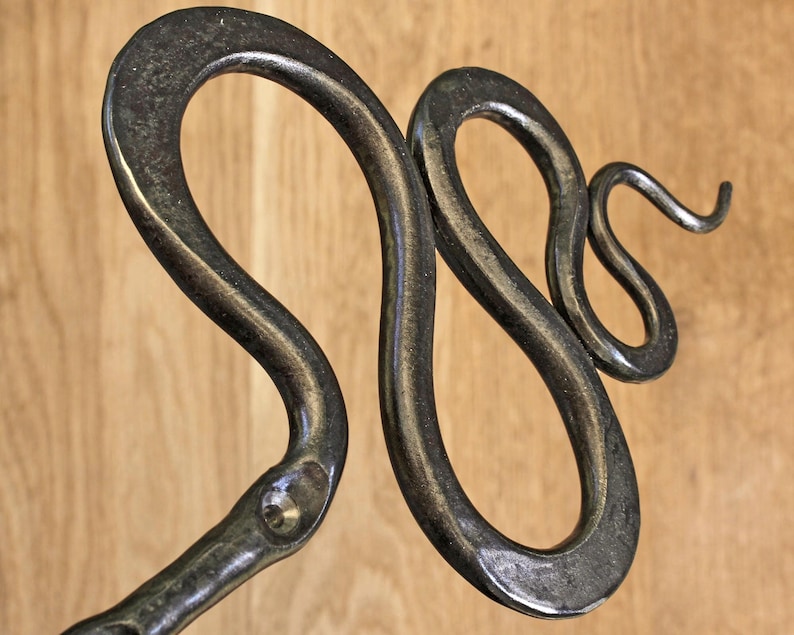 Pu&ograve; includere: Una scultura in metallo forgiato a mano a forma di serpente con una finitura scura e brunita. Il serpente &egrave; raffigurato in una posa sinuosa a forma di S con un corpo liscio e curvo e una testa distinta, dal carattere industriale.