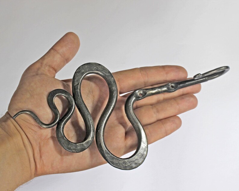 Pu&ograve; includere: Un gancio da parete in metallo forgiato a mano a forma di serpente sinuoso. L'oggetto in ferro grigio scuro e testurizzato presenta un corpo serpentino curvo con due fori di montaggio circolari e un gancio all'estremit&agrave;.