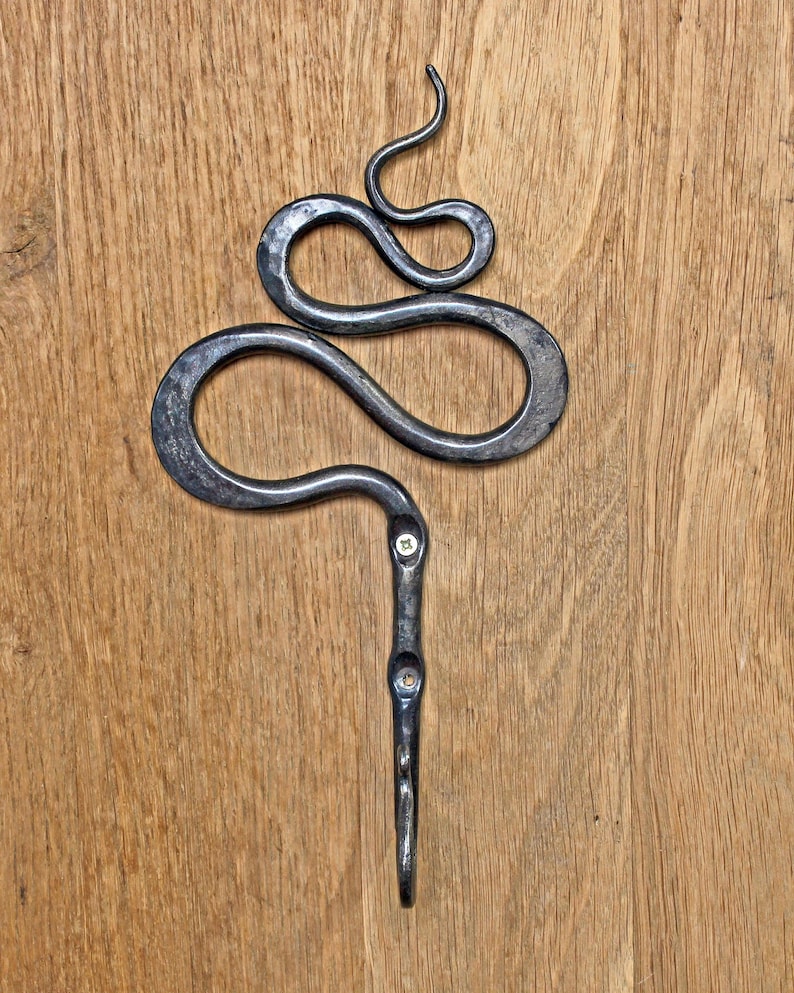 Pu&ograve; includere: Un gancio da parete in metallo grigio scuro forgiato a mano, caratterizzato da un design decorativo a volute che ricorda un serpente nella parte superiore. Il gambo verticale presenta due fori per viti e un gancio curvo.