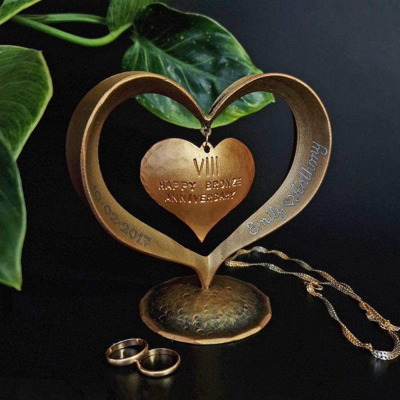 Puede incluir: Una escultura de aniversario en forma de coraz&oacute;n de bronce con las palabras "Happy Bronze Anniversary" y el n&uacute;mero romano VIII. La escultura tiene una base y un colgante de coraz&oacute;n m&aacute;s peque&ntilde;o. Tambi&eacute;n son visibles dos anillos de oro y una cadena de oro.