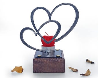 May include: A dark gray metal sculpture featuring two interlocking heart outlines mounted on a rectangular wooden base. A small red apple-shaped candle sits in a silver metal tea light holder between the hearts, with scattered dried petals.
