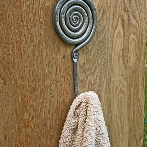 May include: A dark gray metal wall hook featuring a prominent spiral design at the top and a single screw attachment point. A beige textured towel hangs from the curved hook, which is mounted onto a vertical wooden surface with a visible grain.
