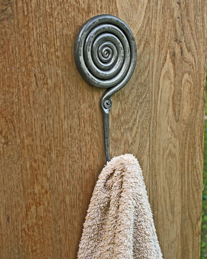 May include: A dark gray metal wall hook featuring a prominent spiral design at the top and a single screw attachment point. A beige textured towel hangs from the curved hook, which is mounted onto a vertical wooden surface with a visible grain.