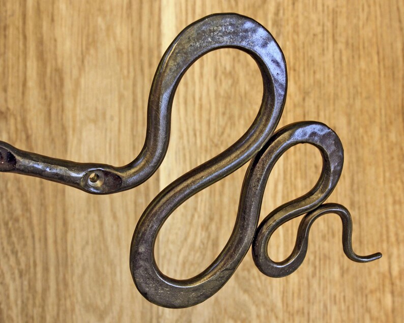 Pu&ograve; includere: Un gancio da parete in metallo martellato scuro a forma di serpente avvolto. Il metallo presenta una finitura testurizzata e usurata con un foro per vite visibile vicino alla testa del serpente. Il corpo si curva in due anelli distinti, terminando con una coda affusolata.