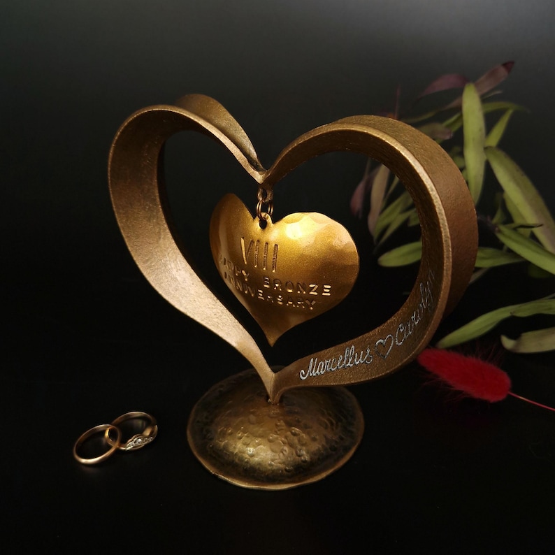 Puede incluir: Una escultura dorada en forma de coraz&oacute;n con un colgante de coraz&oacute;n m&aacute;s peque&ntilde;o, con la inscripci&oacute;n "VIII Bronze Anniversary". La escultura tiene un acabado texturizado y una base. Se ven dos anillos de boda. El fondo es negro.