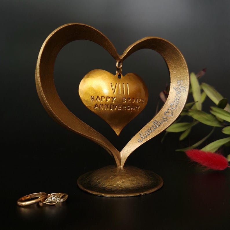 Puede incluir: Una escultura dorada en forma de coraz&oacute;n con un colgante m&aacute;s peque&ntilde;o en forma de coraz&oacute;n con la inscripci&oacute;n "VIII HAPPY BRONZE ANNIVERSARY". Dos anillos de boda de oro con diamantes est&aacute;n en primer plano. La escultura tiene los nombres "Marcella & Candy" grabados.