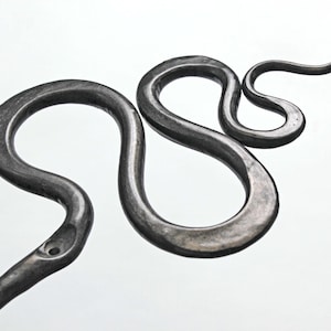 Pu&ograve; includere: Un oggetto in metallo grigio scuro forgiato a mano a forma di figura serpentina sinuosa. La superficie presenta una finitura martellata testurizzata con rientranze visibili lungo il corpo, creando una silhouette a curva a S continua su sfondo bianco.