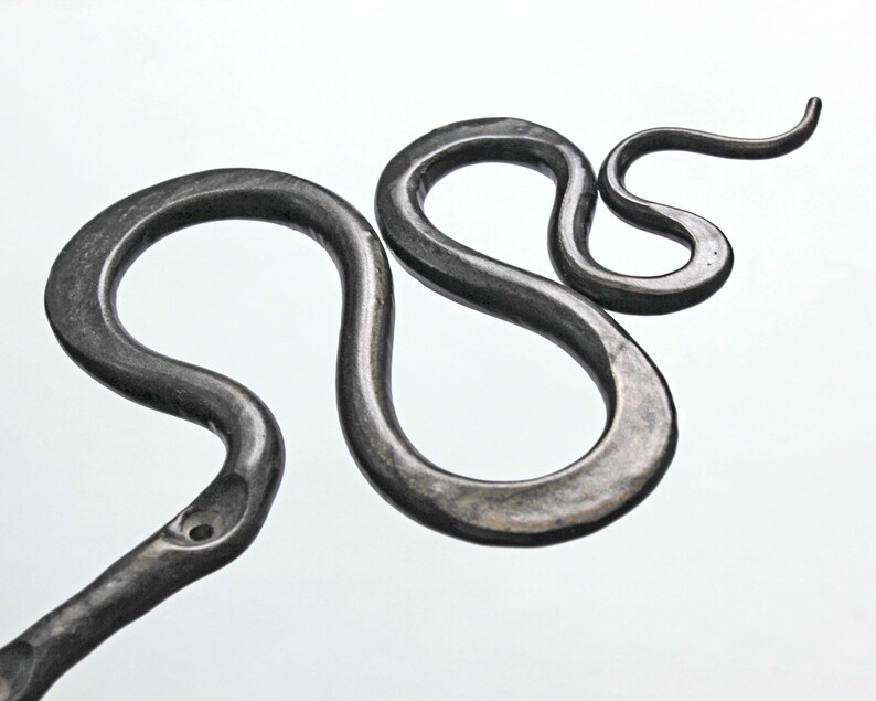 Pu&ograve; includere: Un oggetto in metallo grigio scuro forgiato a mano a forma di figura serpentina sinuosa. La superficie presenta una finitura martellata testurizzata con rientranze visibili lungo il corpo, creando una silhouette a curva a S continua su sfondo bianco.