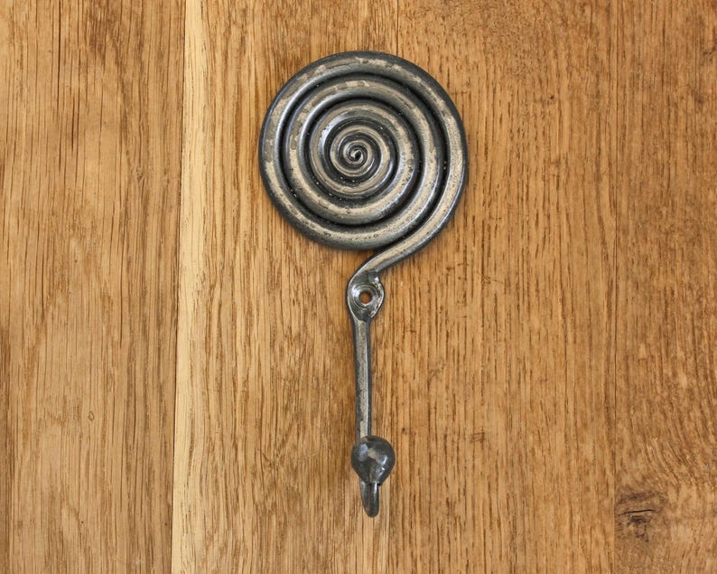 May include: A dark gray metal wall hook featuring a prominent circular spiral design at the top. The hook has a single mounting hole located just below the spiral and terminates in a small, rounded knob at the bottom for hanging items.