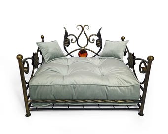 May include: A pet bed featuring a black and bronze-toned metal frame with ornate scrollwork, a central amber-colored glass accent, and a tufted, light sage green velvet cushion. Two matching square velvet pillows rest on the tufted mattress.