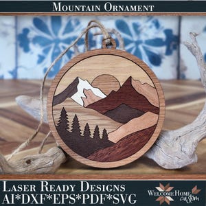 May include: A circular wooden ornament featuring a laser-cut mountain landscape with a sun, layered peaks in varying shades of brown and cream, and a forest of pine trees at the base. The ornament includes a jute twine loop for hanging.