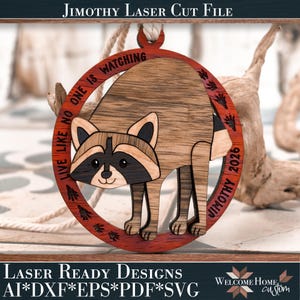 May include: A circular wooden ornament featuring a laser-cut raccoon design. The outer red ring displays the text "LIVE LIKE NO ONE IS WATCHING" and "JIMOTHY 2026" with small paw prints. The raccoon is light brown with dark brown facial markings.