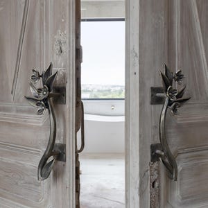 Op de afbeelding: Een paar verweerde, lichtgrijze houten deuren met sierlijke, donkere metalen handgrepen. De handgrepen zijn gevormd als gestileerde bloemen en bladeren. De deuren staan een beetje open en onthullen een helder interieur.