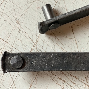 May include: Two black metal drawer pulls with rounded edges and hammered metal texture. Each pull has a single round metal knob.