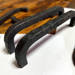 以下が含まれることがあります： ハンマー仕上げの黒い金属製のキャビネットプルが2つ。プルは湾曲しており、シンプルなデザインです。