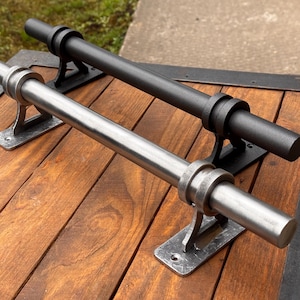 Könnte beinhalten: Zwei Metall-Türgriffe, einer silberfarben und einer schwarz, werden auf einer Holzoberfläche gezeigt. Jeder Griff hat eine zylindrische Stange, die von dekorativen Halterungen gehalten wird. Das Holz hat einen warmen, rötlich-braunen Ton.