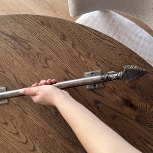 May include: A silver metal arrowhead door handle with a decorative design. The handle is mounted on a wooden surface.