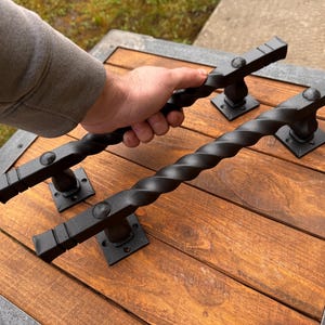 May include: Two black metal door handles with a twisted design. The handles are mounted on a wooden surface.