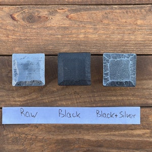 May include: Three square-shaped stone tiles in different finishes: raw, black, and black with silver accents. The tiles are arranged horizontally on a wooden surface with a blue paper label underneath that reads "Raw", "Black", and "Black + Silver".