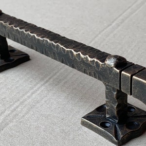 May include: A rustic, hammered iron door pull with a square design. The handle is a dark bronze color and has a textured finish.