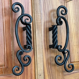 Puede incluir: Par de manijas de puerta de hierro forjado negro con un diseño decorativo de volutas. Las manijas tienen un agarre central retorcido y están montadas en una puerta de madera. Las manijas miden aproximadamente 25 cm de largo.