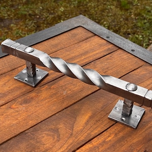 Op de afbeelding: Een gedraaide metalen deurgreep met vierkante uiteinden en een zilveren afwerking. De greep is gemonteerd op een houten deur met een zwart metalen frame.