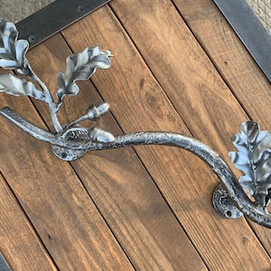 Puede incluir: Un perchero de metal plateado con forma de rama de roble con hojas y bellotas. El perchero está montado en una pared de madera.