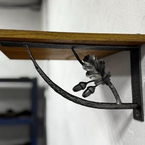 Op de afbeelding: Een houten plank met zwarte metalen beugels met een eikenblad- en eikelontwerp. De beugels zijn gebogen en hebben een rustieke, handgemaakte uitstraling.