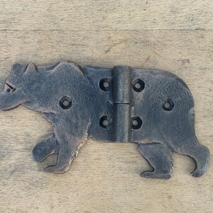 May include: A decorative metal hinge in the shape of a bear, with a dark gray finish. The bear is depicted in a walking pose, with visible details like the ears, paws, and facial features. The hinge has multiple screw holes.