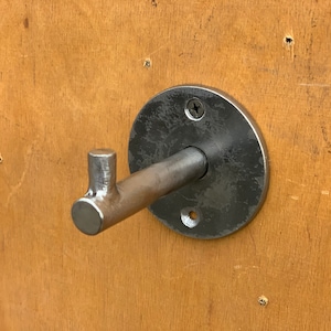 May include: A silver metal hook mounted on a round metal plate. The hook is attached to a wall with a screw.