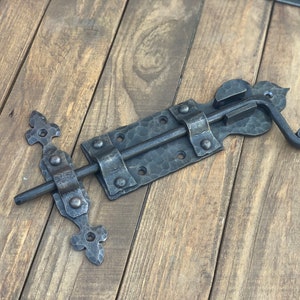 May include: A black metal door latch with a decorative scroll design. The latch has a hammered finish and is mounted on a wooden surface.