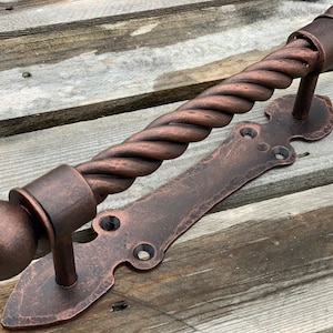 May include: A twisted copper door handle with a decorative plate and rounded ends. The handle has a rustic, aged finish.