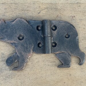 May include: A rustic, metal hinge in the shape of a bear. The hinge is dark brown with visible texture and features multiple screw holes. The bear is depicted in a walking pose, adding a unique decorative element.