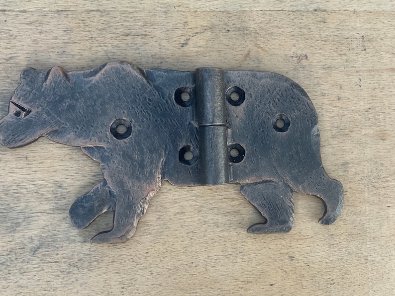 May include: A rustic, metal hinge in the shape of a bear. The hinge is dark brown with visible texture and features multiple screw holes. The bear is depicted in a walking pose, adding a unique decorative element.