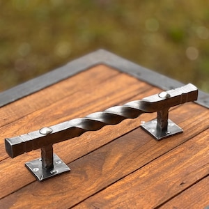 Op de afbeelding: Een gedraaide metalen handgreep met vierkante uiteinden en een zilveren afwerking. De handgreep is gemonteerd op een houten oppervlak met een zwarte rand.