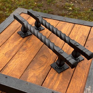May include: Two black twisted metal door handles with square bases mounted on a wooden surface.